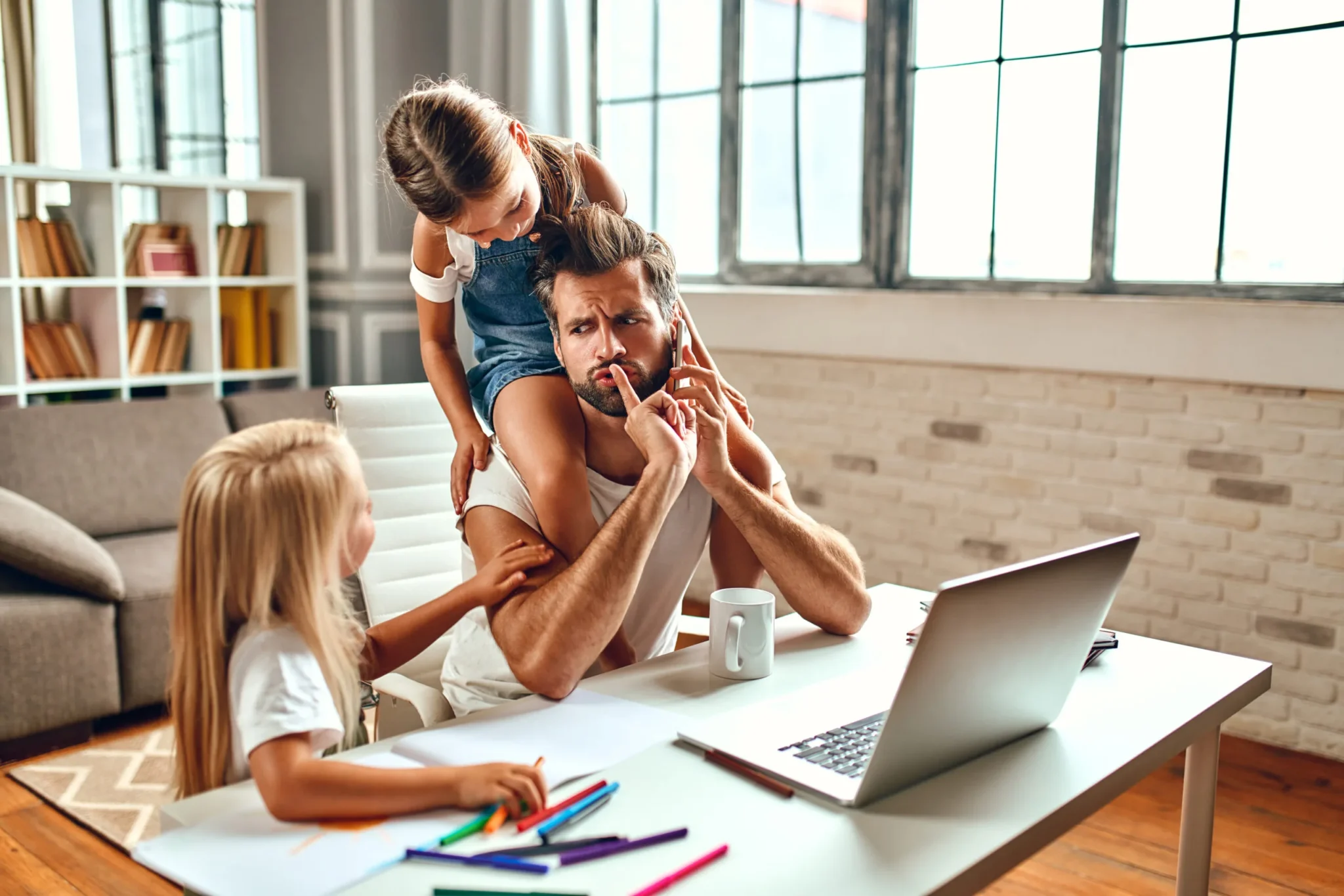 The businessman dad tries to work at the laptop when his little daughters play, fool around and interfere with him. Freelance, work from home.